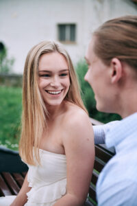 A young woman in a white dress smiles as she talks with a young man in a blue shirt, both seated on a park bench near a white building.