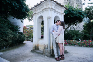 A couple shares a kiss in front of a small, white, arched structure in a garden. The man wears light-colored pants and a shirt, while the woman wears a white dress and black shoes. Surrounding greenery and colorful flowers enhance the romantic atmosphere.