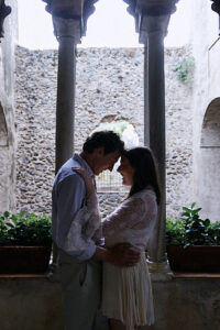 A couple stands close together, foreheads touching, in a stone archway with plants on either side. They are embracing, staring into each other's eyes lovingly. The background features a stone wall and an overcast sky, giving a romantic and intimate atmosphere.